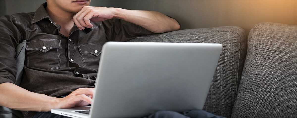 Man sitting on his couch. He is using a laptop and is clearly thinking deeply.